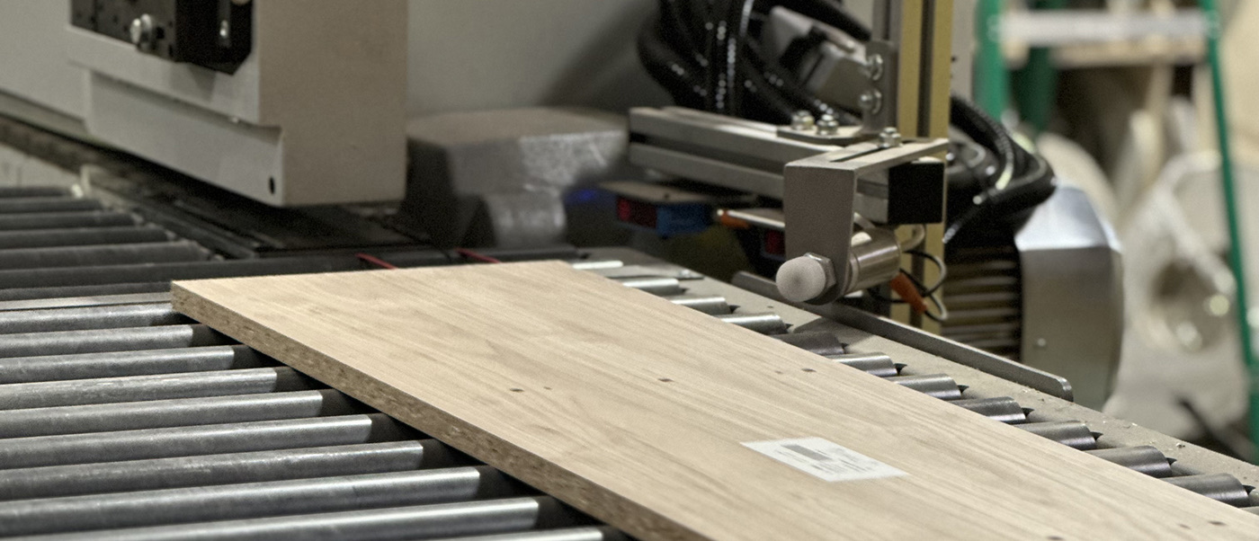 Oak cabinet panel moving on conveyor rollers toward woodworking machinery in cabinet production line in Ronbow Livermore California factory.